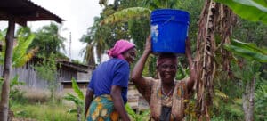 Two women outside laughing while one carries a blue bucket on her head.