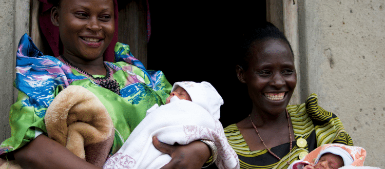 Two women stand outside and smiling as they hold their babies.