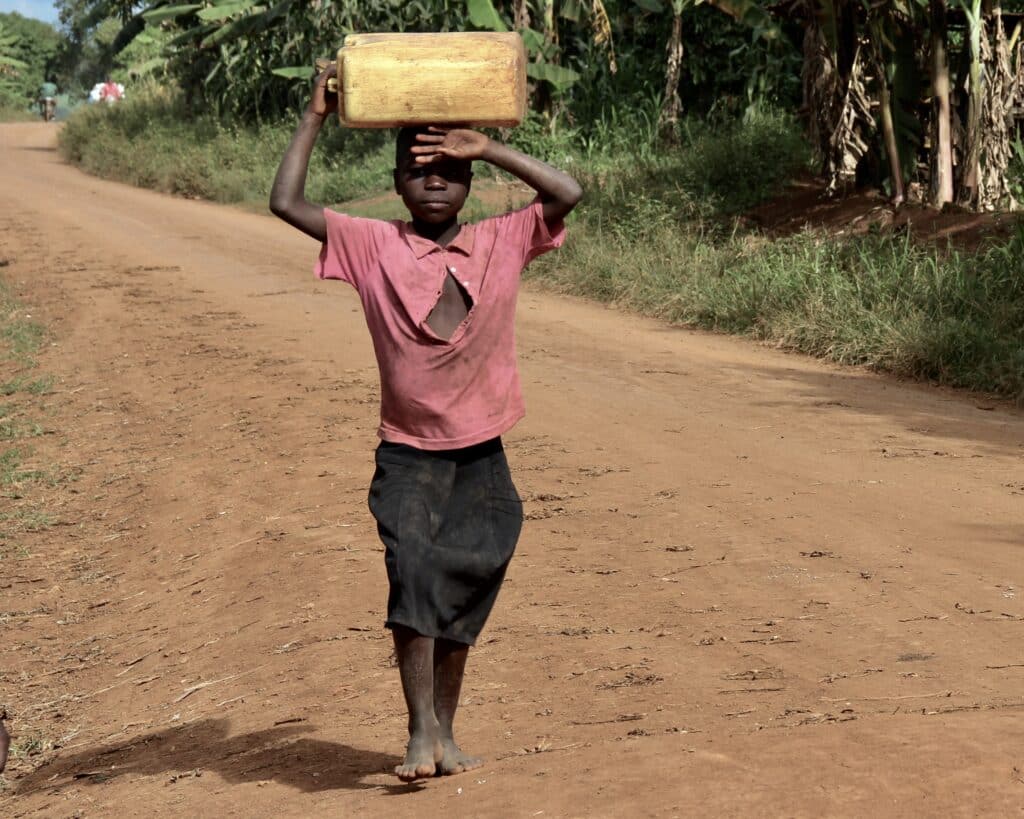 Boy walking on dusty road holding a water jug on his head.