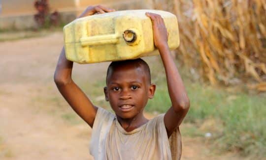 Boy on dusty road holding water jug on his head.