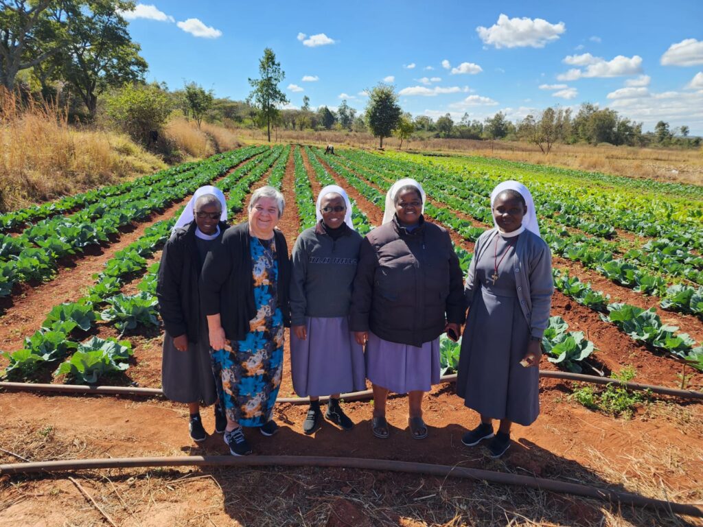 Five women smiling in a front of crops grown in neat rows.