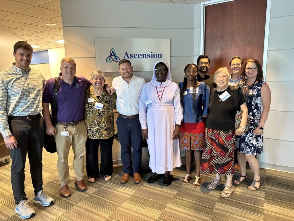 A group people smiling in front of a sign with the "Ascension" logo.